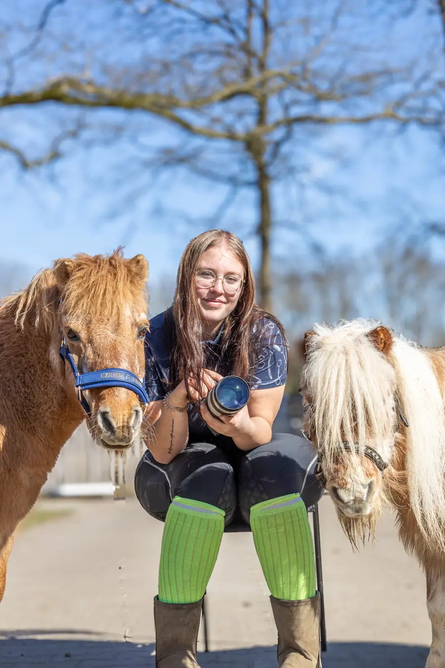 Een media stagiair zit tussen twee Shetlandpony’s en houdt een camera met telelens vast. De pony links draagt een blauw halster, terwijl de pony rechts een lange, lichte manen heeft. Op de achtergrond zijn een boom en een heldere blauwe lucht te zien.