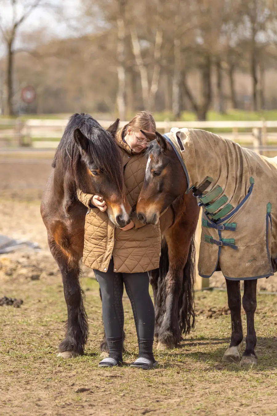 Persoon staat tussen twee paarden in een weiland en knuffelt ze liefdevol, terwijl de paarden hun hoofden tegen elkaar en de persoon aan houden.