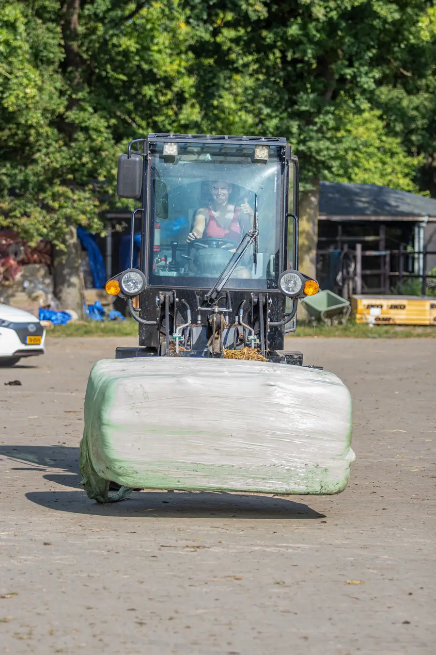 Een vrijwilliger bestuurt een shovel over het terrein van de paardenopvang en vervoert een ingepakte hooibaal. Vanuit de cabine steekt de bestuurder lachend een duim omhoog, terwijl op de achtergrond stallen en bomen zichtbaar zijn.